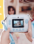 Close-up of a child holding the kiimento Model P Cinnamoroll camera, showing a preview screen with a girl posing for a photo. The lanyard strap with Cinnamoroll branding is visible, set against a cheerful background with illustrated accents.