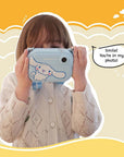 A young girl holds up the kiimento Model P Cinnamoroll kids’ digital camera as if taking a photo, with playful text reading “Smile! You’re in my photo!” on a cheerful yellow background with cartoon clouds.