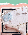 Lifestyle image of the kiimento Model P Cinnamoroll kids’ digital camera placed on an open scrapbook filled with printed photos. A child's hand is seen writing in the book, capturing a sweet moment of creativity and memory-keeping.