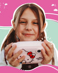 Smiling young girl holding the kiimento Hello Kitty Model P digital camera, showing joy and excitement. The front of the pastel pink camera with Hello Kitty’s face is clearly visible. The background features playful illustrations in pink, mint green, and white, evoking a cheerful, whimsical atmosphere.