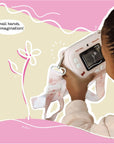 Close-up of a young girl using the Kidamento Hello Kitty Model P camera to take a photo, viewed from behind. The camera screen displays a flower sketch, and the camera strap with Hello Kitty designs hangs around her hands. Caption reads “Small hands. Big imagination!” over a soft pink and beige illustrated background.