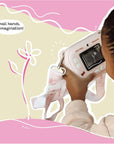 Close-up of a young girl using the kiimento Hello Kitty Model P camera to take a photo, viewed from behind. The camera screen displays a flower sketch, and the camera strap with Hello Kitty designs hangs around her hands. Caption reads “Small hands. Big imagination!” over a soft pink and beige illustrated background.