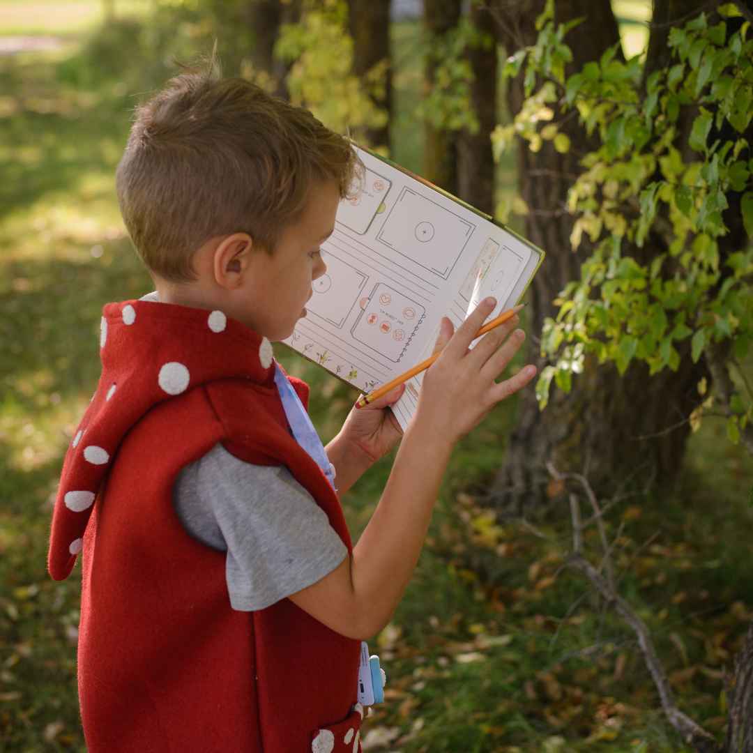 A child in a red mushroom-themed vest holds an activity book and pencil outdoors.