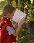 A child in a red mushroom-themed vest holds an activity book and pencil outdoors.
