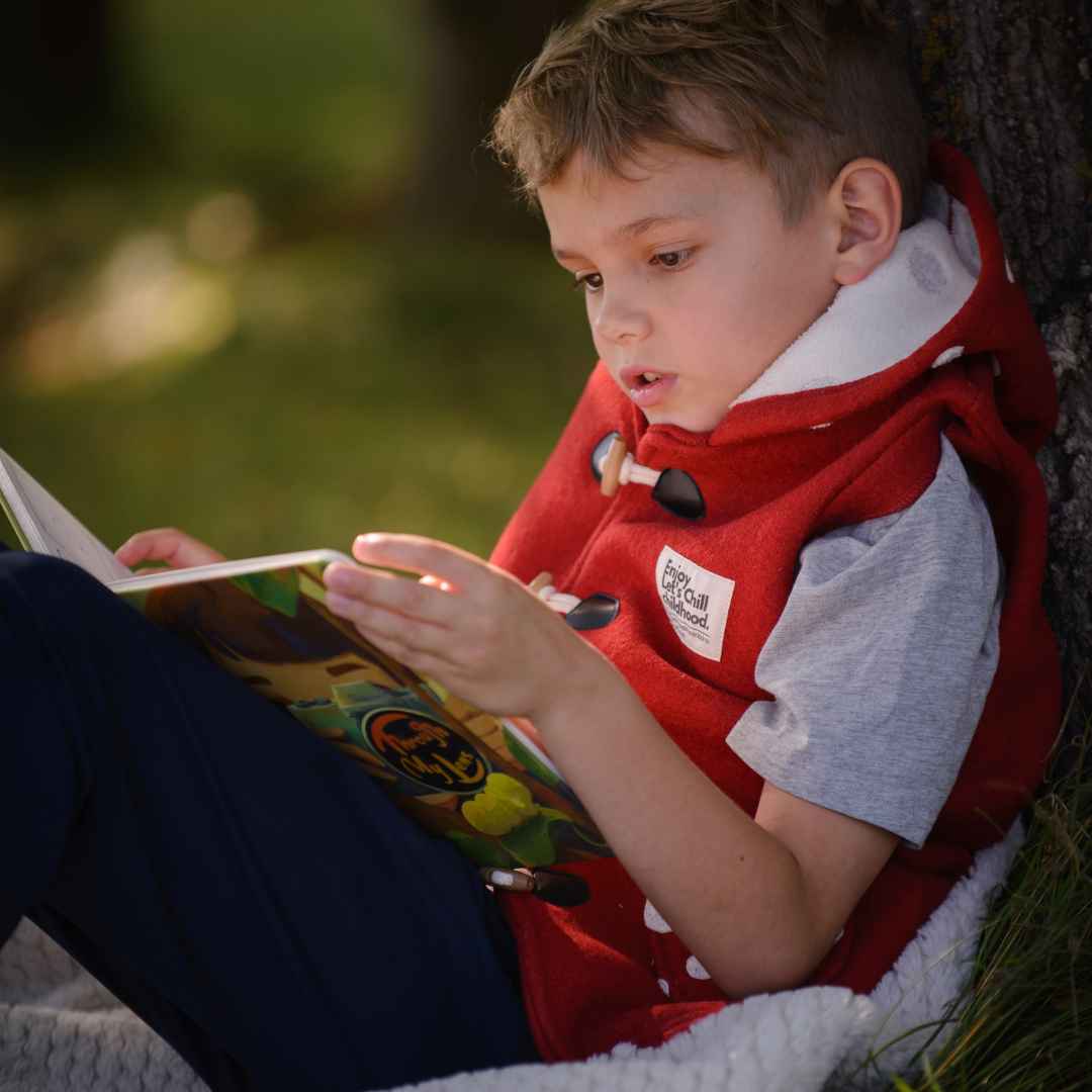 A young boy in a red vest reads an activity book outdoors.