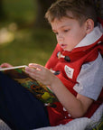 A young boy in a red vest reads an activity book outdoors.