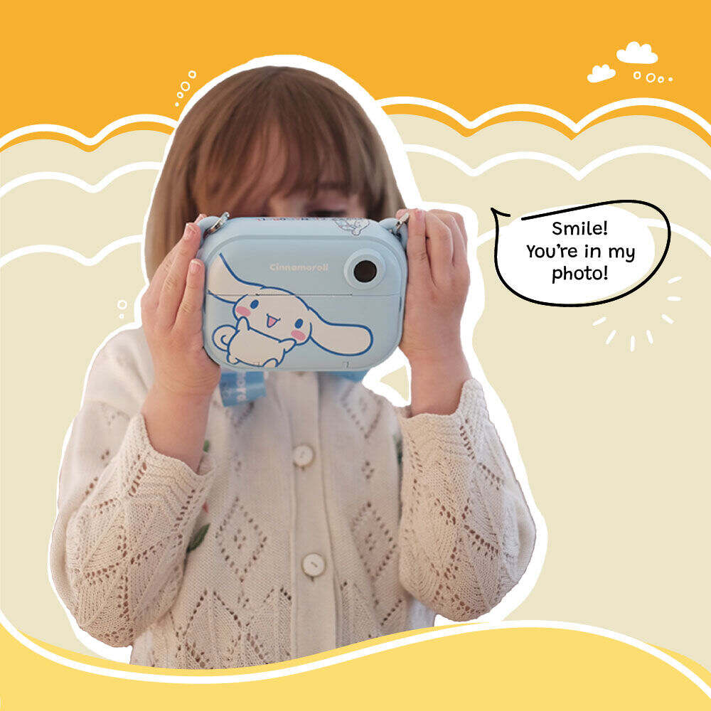 A young girl holds up the Kidamento Model P Cinnamoroll kids’ digital camera as if taking a photo, with playful text reading “Smile! You’re in my photo!” on a cheerful yellow background with cartoon clouds.