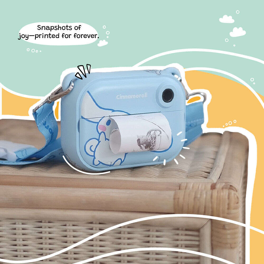 Kidamento Model P Cinnamoroll kids’ digital camera placed on a wicker table, actively printing a black-and-white photo. The scene is framed by a colorful illustrated background with the caption “Snapshots of joy—printed for forever.”