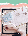 Lifestyle image of the Kidamento Model P Cinnamoroll kids’ digital camera placed on an open scrapbook filled with printed photos. A child's hand is seen writing in the book, capturing a sweet moment of creativity and memory-keeping.