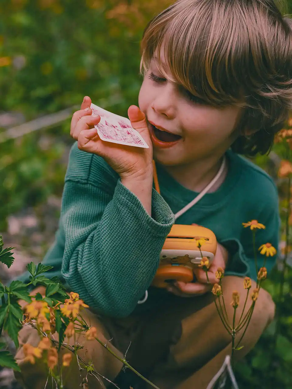 Child holding a small toy camera and a pink flower in a natural setting with greenery.