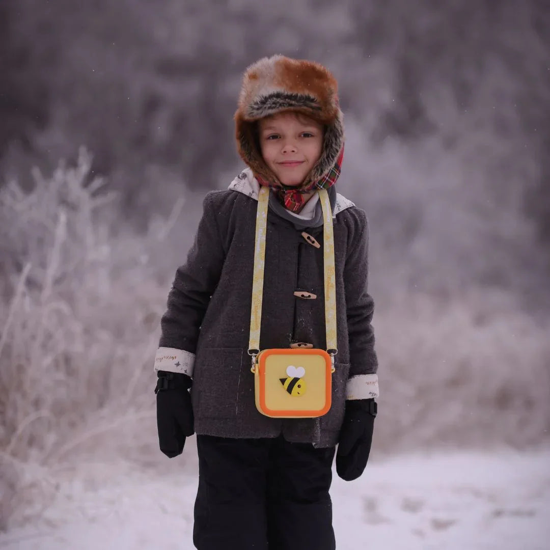 A child wears a grey coat and a fur hat, with a yellow bee-themed silicone bag.