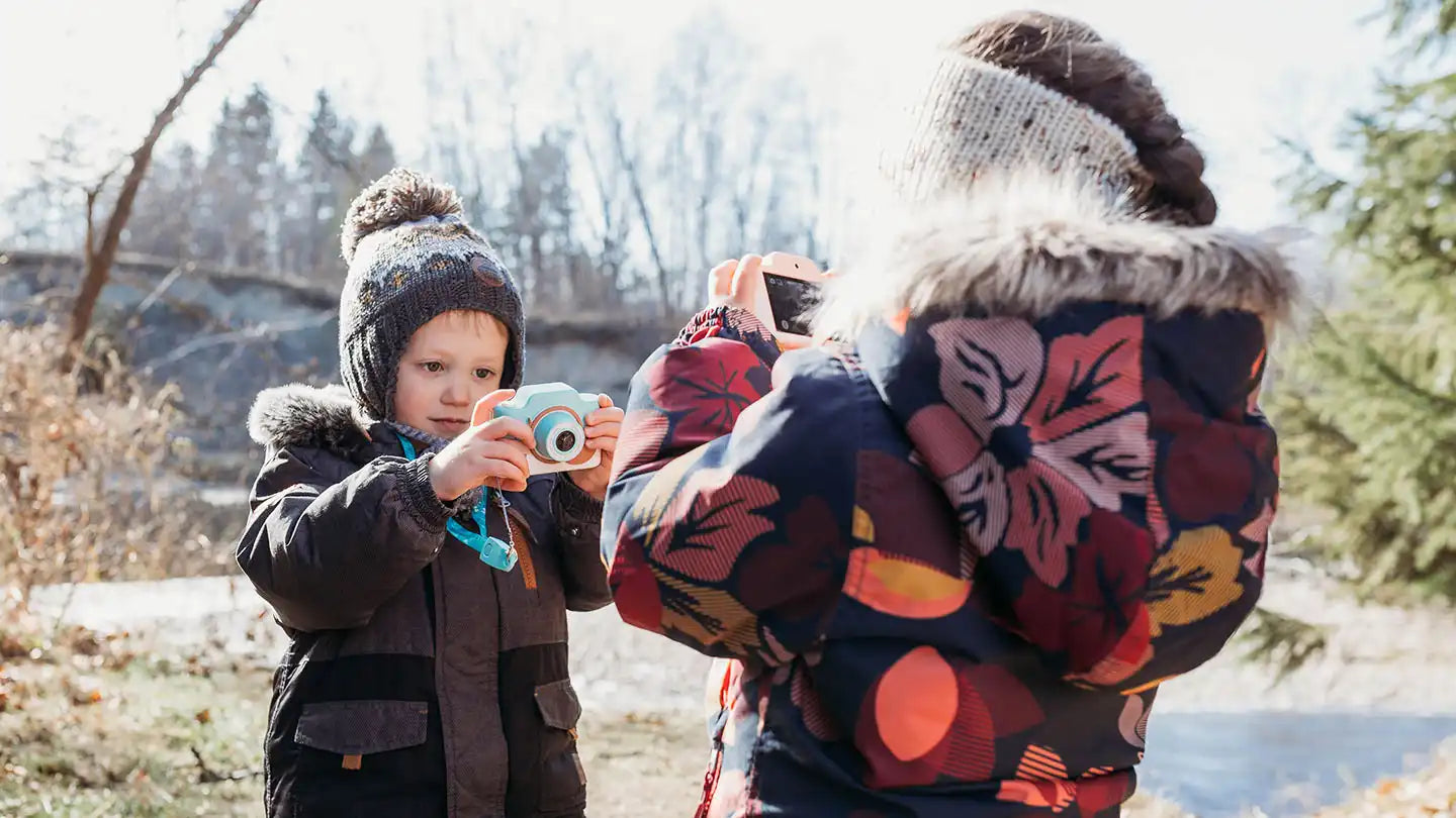 Child taking a photo of an adult with a camera outdoors in winter.